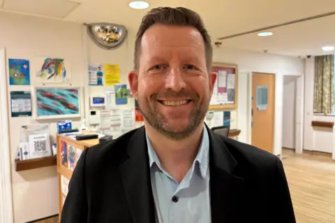 Man with short brown hair, short beard and wearing a black blazer, light blue shirt smiles at the camera. In the background is office space, and pictures on the back wall, and wooden flooring.