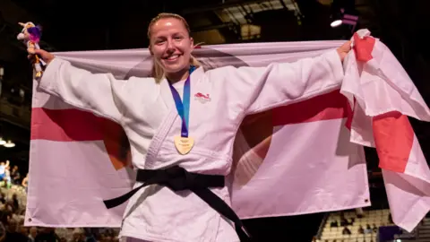 Kelly is wearing a white judo kit with a gold medal around her neck and black belt. She is holding an England flag behind her.
