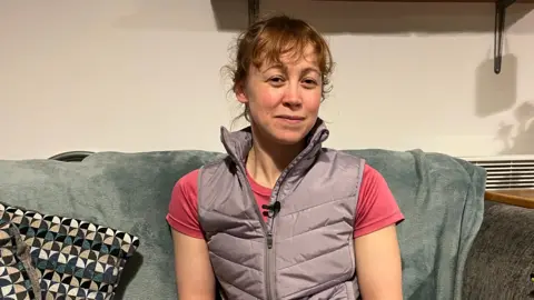 A woman in a light purple gilet and pink T-shirt sat on a sofa which has a grey blanket over it. To the left of her is a patterned cushion with green, white, cream and black patterns on a grey background. Behind her is a radiator to the righter and brackets holding up a shelf just out of shot.