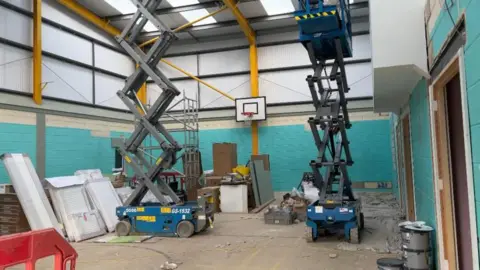 An indoor basketball court, with blue walls, filled with building equipment, it is being renovated with two blue electric lifts in the centre.