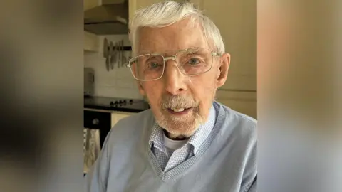 World War Two and D-Day veteran Tom Berry, 100, has grey hair and is wearing silver glasses, a white vest, grey checked shirt and grey v-neck jumper. Mr Berry is sitting in a kitchen.