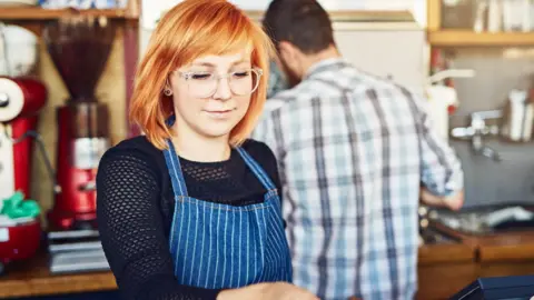 A young woman with strawberry blonde hair and clear-rimmed glasses working behind a counter in a coffee shop - she is wearing a black long-sleeve top and a blue pin-striped apron. Behind her is a man in a white checked shirt with his back to the camera.