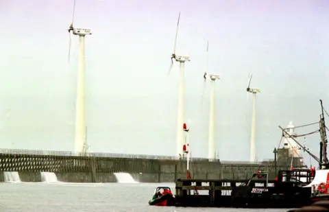 PA Media A photo of the wind turbines being built in Blyth. There is a large concrete structure supporting four turbines. A black vessel with the name "Bernica Newcastle" is next to it.