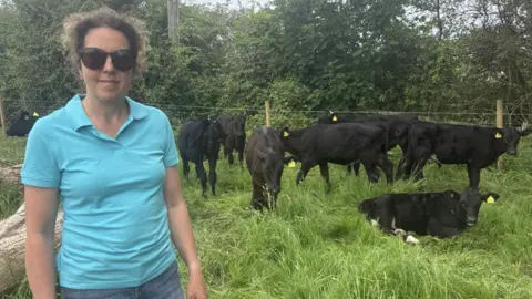 Leonie McIntosh A woman in a blue top with sunglasses. She has brown curly hair. She is stood outside in front of a number of black cows in a grassy field. There are trees behind her. 