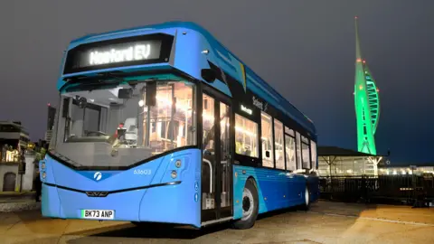 Promotional photograph of First Bus Solent electric vehicle. The single decker bus is light blue. In the background, the Spinnaker Tower is illuminated green.