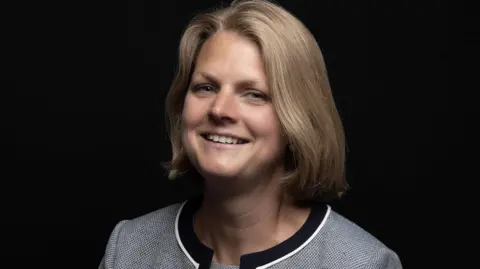 Abi McGuire Abi McGuire smiling in a grey suit against a black screen