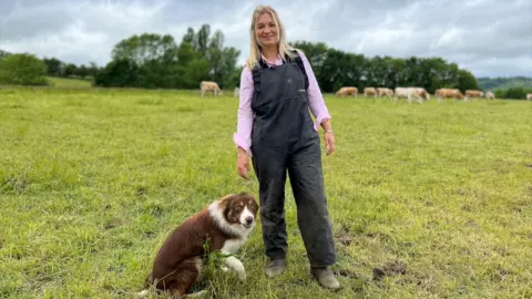 Harriet Cowan stands in a field with a dog, wearing green overalls. There are cows in the background