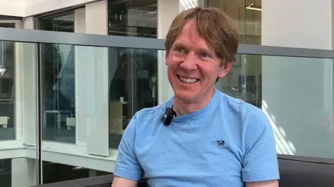 Peter Bell smiling widely and happily while looking to the side - he is wearing a light blue shirt and has blonde mid-length hair.