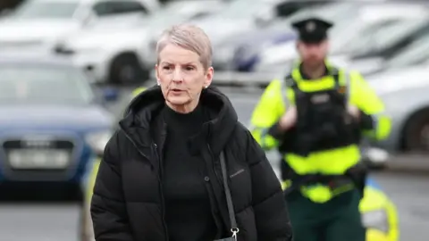 PA Media Eleanor Donaldson con un largo abrigo negro, top negro y con un bolso negro. Ella está mirando fuera de la cámara. En segundo plano, un oficial de policía uniforme y algunos autos fuera de foco.