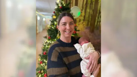 Chloe and her newborn son Alfie pictured in front of a Christmas tree, a short time after his birth on Christmas Day 2024. Chloe is wearing a blue and brown striped jumper and is holding Alfie on her left side. Alfie is wearing a cream Babygro. 