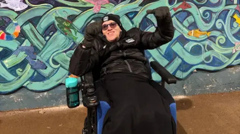 A man wearing a black jacket and hat, sitting in a wheelchair. He has his hands raised in the air.