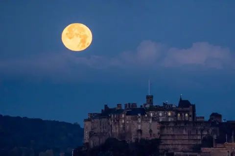 Jane Barlow/PA Media The full October moon, known as the Hunter's sets behind Edinburgh Castle, Scotland. 17 October 2024. The moon is on the left hand side of a blue dusk sky. The castle in in the foreground to the right.