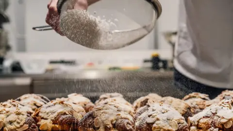 Thomas Weller Photography A baker sprinkling powder sugar over almond croissants.