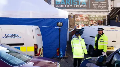 John Fairhall/BBC Police officers outside a building with a sign which reads BARISTA. There is a blue and white police tent, a police cordon and a police car which reads FORENSIC INVESTIGATION.