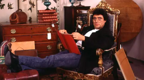 Actor Ian McShane wearing jeans, boots, a black jacket, glasses, and a white T-shirt. He is sitting on a throne-like chair with his legs perched on a red leather stool. He is holding a red book and looking directly into the camera. 