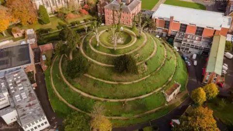 Marlborough College/Peter Davies Photography An aerial view of the Marlborough Mound. It is a grassy hill with six rings cut into its side. It has a few trees on top and is surrounded by buildings.