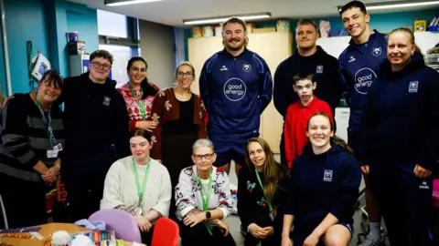 A group of about a dozen people pose for a photo with just over half of them stood behind while four women kneel in front. They are of varying ages and a mix of men and women. Some of them are from Bristol Bears.