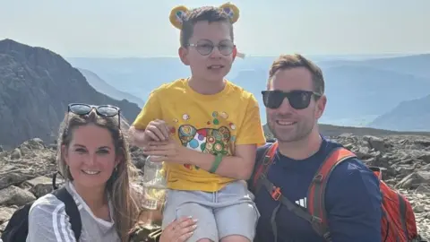 Family photo A boy with light brown hair and glasses with a yellow tshirt and yellow bear ears being held up my a man with brown hair, sunglasses and a blue tshirt and a woman with blonde hair and sunglasses on top of her head wearing a grey tshirt, all stood on rocky ground