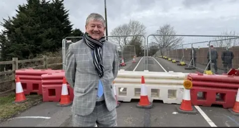 Staffordshire County Council A man with short grey hair, wearing a blue and white striped scarf, and a grey checked suit (with blue tie peeping out) stands on a road in front of bollards showing the road is closed