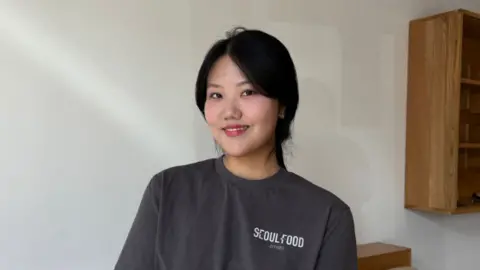 Jae Shon, Seoul Food + Studio An image of a smiling young woman with black hair tied back wearing a dark grey t-shirt that says "Seoul Food" in white writing. She is sitting on a beige sofa with wooden backing. The wall behind her is white and there are wooden shelves on the wall to the right.