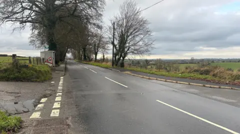 A wide view of a rural crossroad junction outside Rathfriland town. There is a grey bus shelter on the left, close to the crossroads, and a red roadworks sign rests close by on the grass verge. The road beyond is lined with a tunnel of trees. Green fields stretch out to the horizon on either side of the road.