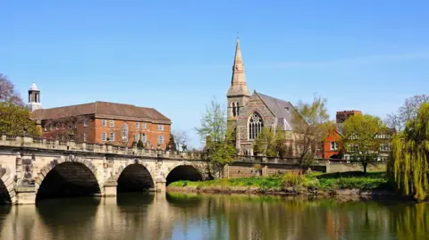 An arched stone bridge over a river with a church spire and a large brick building seen on the far side and the buildings and trees reflected in the water