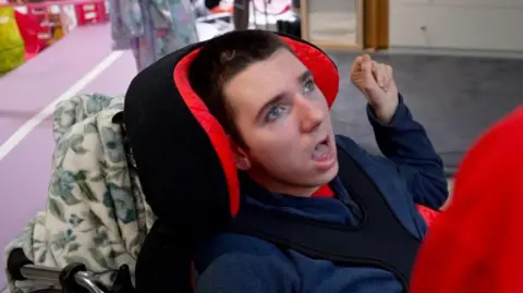 A boy in a chair with a head support holds his hand in the air. There is a floral blanket behind him. 