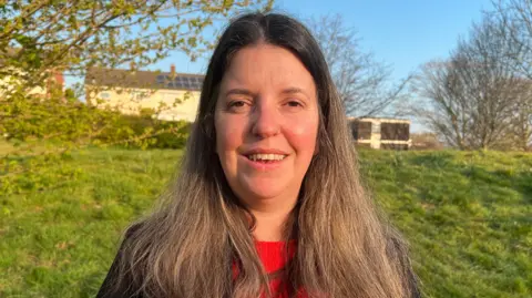 Federica Smith-Roberts is pictured standing in front of a hill. She is smiling directly at the camera and she is wearing a red jumper underneath a black jacket. The tops of some houses can be seen just beyond the hill behind her.