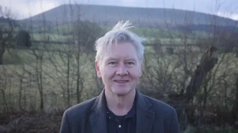 A man with grey hair and wearing a dark jacket is outside smiling at the camera. Green fields, a hill and the sky are in the background.  
