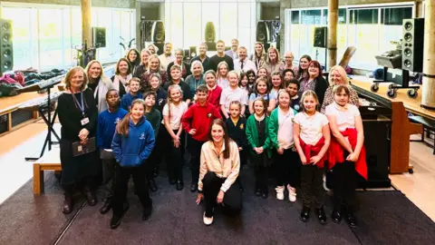 A group of primary school pupils smiling at the camera,wearing their uniform jumpers and hoodies gathered together with their teachers in the middle of a recording studio. You can see speakers and recording equipment round them