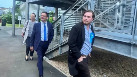 Three people including defeated MP Steve Baker walk along a paved path beside a metal stair structure and industrial-style building, led by a person in a blue suit and tie, with another in a dark coat wearing a blue rosette badge walking slightly ahead; a third person follows behind.