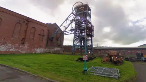 A red brick building with arched windows. To the right of the building is a large metal structure. In the foreground, there is a green post with a sign reading Haig Mining Museum.