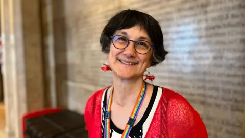 A head and shoulders image of a woman with short dark hair. She has round glasses and is wearing a rainbow lanyard, a red cardigan and a white and black top.
