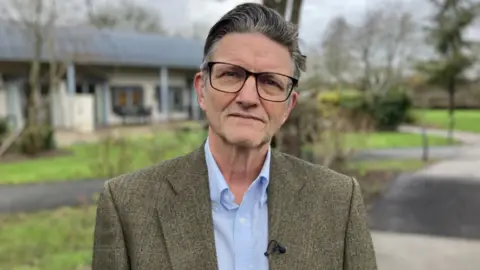 A man wearing glasses, a tweed jacket and glasses stands outside with grass and trees behind him, and a building behind that.