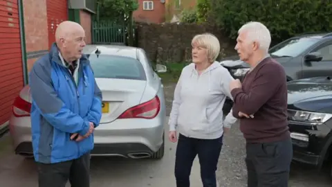 A man talks to a man and a woman who are stood opposite him. There are three parked cars in the background as they are outside a car repair garage. 