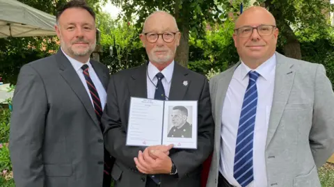 BBC/Adele Forrest Three men stood in suits in front of a memorial plaque. The man in the centre holds a black and white photograph of his father.