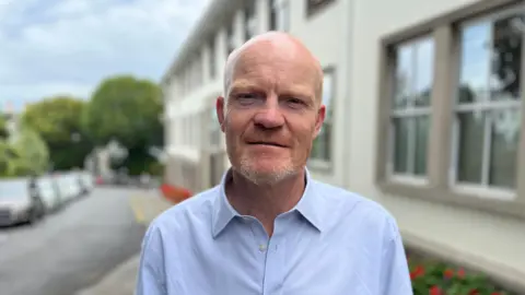 Deputy Gavin St Pier - A bald man with a ginger and grey speckled beard. He's wearing a blue buttoned shirt with the collar undone. 