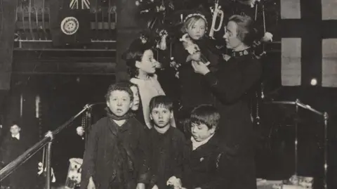 Salvation Army A black and white photo of a woman in Salvation Army uniform holding a child. She is beside four other children who are looking at the camera. 