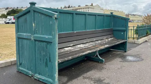 A large brown wooden public bench with green panelling around it. It is on a tarmac walkway with grass behind it