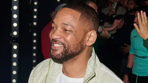 Will Smith at Baller League in London. He has a short beard and cropped hair and wears a pale green jumper over a white T-shirt. He smiles, looking away from the camera, with crowds seen behind him in the background. 