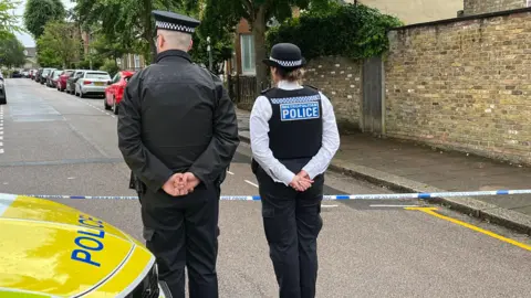 Two Metropolitan Police officers standing with their backs to the camera behind police cordon tape on a residential street.