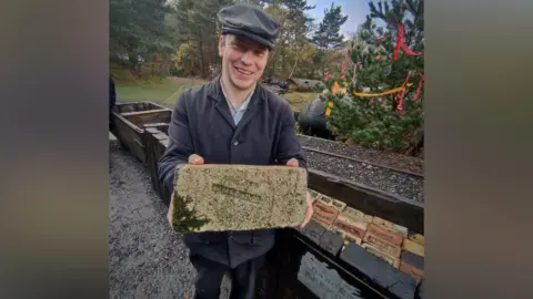Danijel Borna Fiket Alexander Appleton is smiling as he holds a brick while standing in front of a narrow brick path at Beamish Museum. He is wearing a black buttoned-up shirt and black trousers, as well as a dark grey baker boy hat. 
