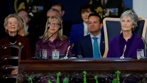 PA Media Catherine Connolly with short grey hair, wearing a purple top and purple jacket, sits at an ornate table. In the background are Mary Robinson, who has short grey hair and is wearing a purple jacket over a black jumper; Mary McAleese who has shoulder length blonde hair, glasses and is wearing a purple dress; and Leo Varadkar who has short black hair and wearing a dark blue jacket and a blue shirt and tie.