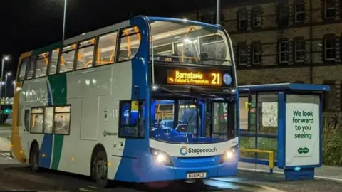 A double-decker bus, pictured at night at a deserted bus stop. Operated by Stagecoach, the bus is painted blue, white, green and yellow and has  "Barnstaple" on its destination board.