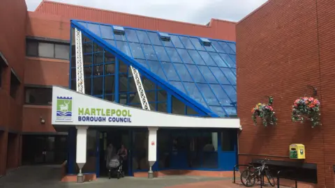 The outside of Hartlepool Borough Council which is a large, red brick building with an angled glass entrance with blue window panes. Hartlepool Borough Council is written in green and blue above the entrance and there are two people pushing a pram underneath.