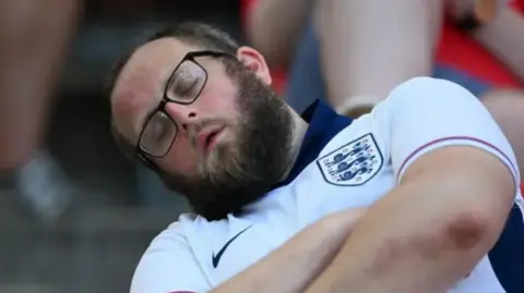 Getty Images A man asleep. He is wearing glasses and a white England shirt. 