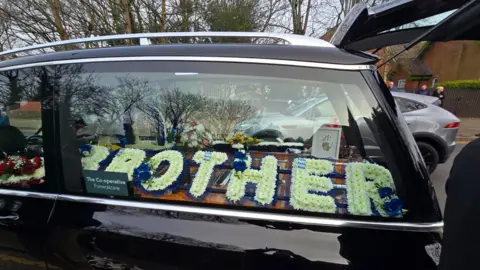 A close-up on the window of a black hearse showing white and dark blue flowers arranged to say 'brother'. A small framed purple handprint is also visible.