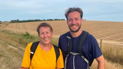 Alex and Miles Eades are looking at the camera. Alex is wearing a yellow top, while Miles is in blue. They are wearing backpacks. Behind them are yellow fields. Their faces are red from the heat and they are smiling.