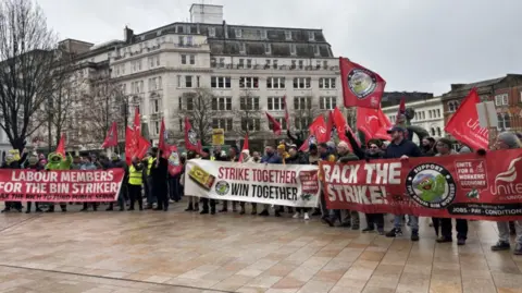 Dozens of protesters holding red unite the union flags and three large banners with messages in support of the bin strikes.