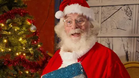 Ade Bowen A man is dressed as Santa, with a red hat and red coat. He has white long hair and a white beard and is smiling at the camera. He is holding a blue, glittery present and a Christmas tree can be seen behind him.